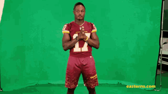 a football player is standing in front of a green screen wearing a red jersey and shorts .