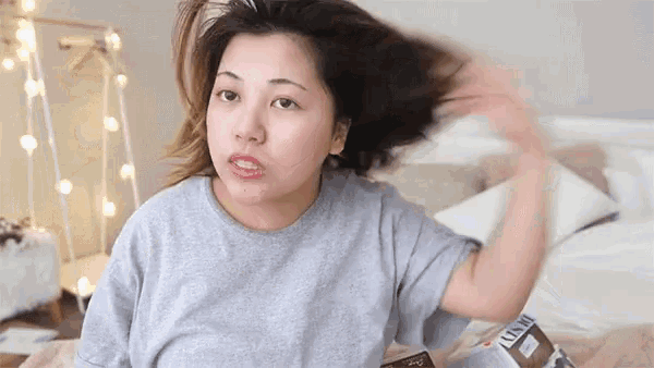 a woman in a grey t-shirt is sitting on a bed with her hair blowing in the wind