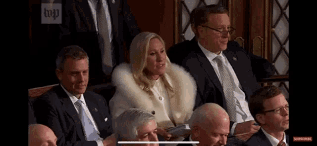 a woman in a white fur coat is sitting in a row of men in suits .