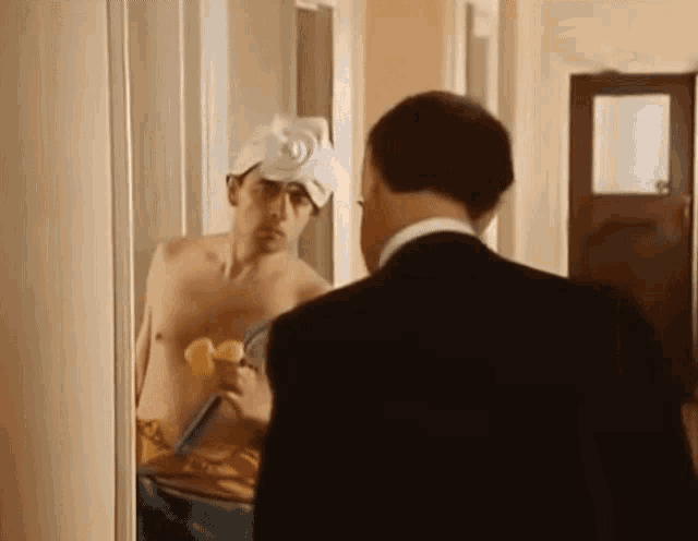 a man in a suit looks at a shirtless man in a towel on his head