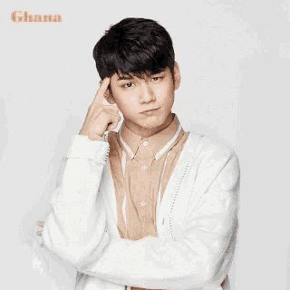 a young man wearing a white sweater and a tan shirt is holding his finger to his forehead