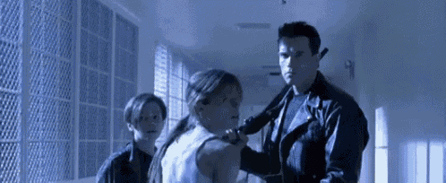a man in a leather jacket is holding a gun in a hallway while a girl looks on .