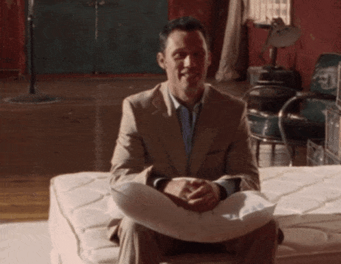 a man in a suit sits on a mattress with a pillow