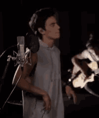 a man in a white shirt is standing in front of a microphone while another man plays a guitar .