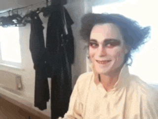 a man with makeup on his face is smiling in a room with clothes hanging on a rack .