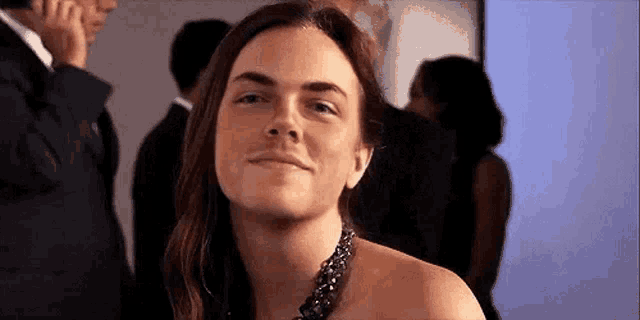 a woman with long hair and a necklace is smiling at the camera while standing in a crowd .