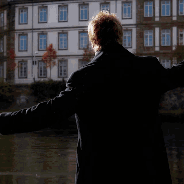 a man in a black coat is standing in front of a building with his arms outstretched