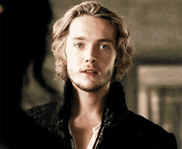 a young man with long blonde hair and a beard is wearing a black jacket and white shirt .