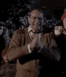 a man in a brown sweater is standing in front of a group of people and clapping his hands .