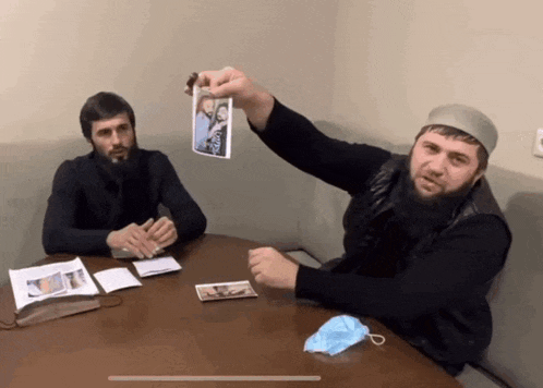two men sitting at a table one of whom is holding up a picture of a man with the word russia on it