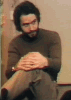 a man with a beard wearing a brown sweater sits with his legs crossed