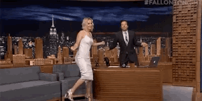 a woman in a white dress is jumping in front of a man in a suit on a talk show .