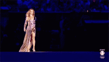 a woman in a long dress is walking on a stage with the olympics logo in the background