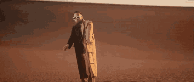 a man wearing sunglasses and a trench coat stands in the desert
