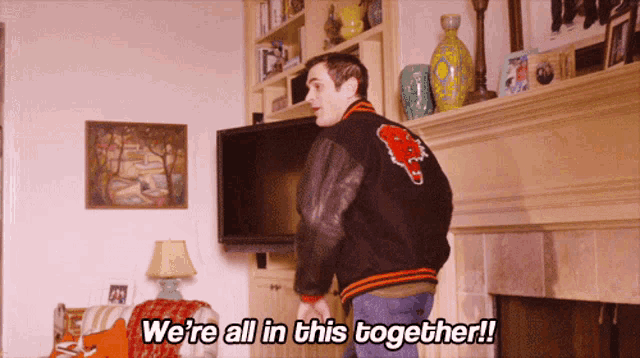 a man in a varsity jacket is standing in a living room and says we 're all in this together