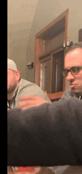 two men are sitting at a table with one wearing glasses and making a funny face .