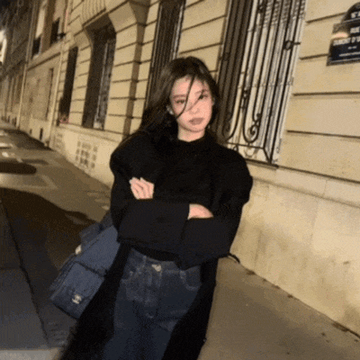 a woman wearing a black sweater and jeans stands in front of a building with a street sign that says rue de la paix
