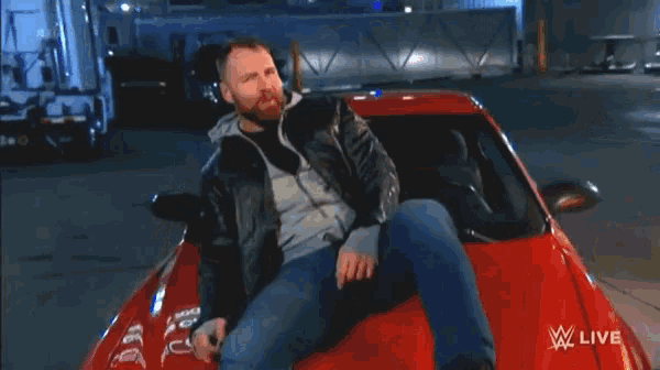 a man in a leather jacket stands in front of a red car that is on fire and says wwe live on the bottom