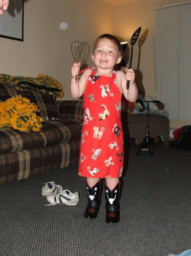 a young boy in a red apron and cowboy boots holds a spatula