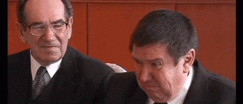 two men in suits and ties are sitting next to each other and one is making a funny face