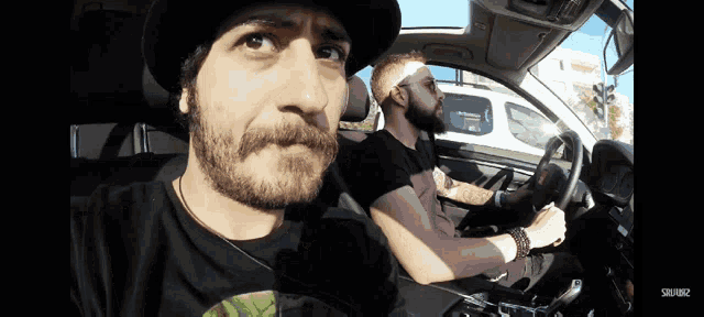 a man with a beard is driving a car with another man in the back seat