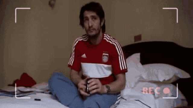 a man in a red adidas shirt sits on a bed in front of a camera