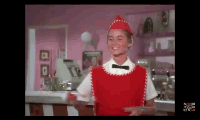 a woman in a red apron and red hat is standing in front of a pink counter .