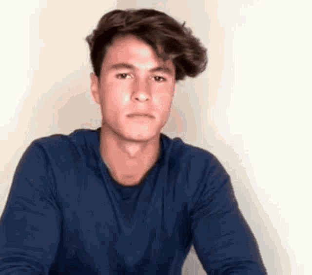 a young man in a blue shirt is sitting in front of a white wall and looking at the camera .