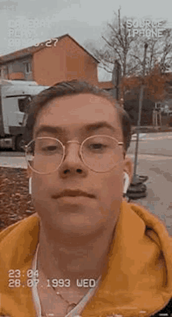 a young man wearing glasses and ear buds is taking a selfie with a camera .