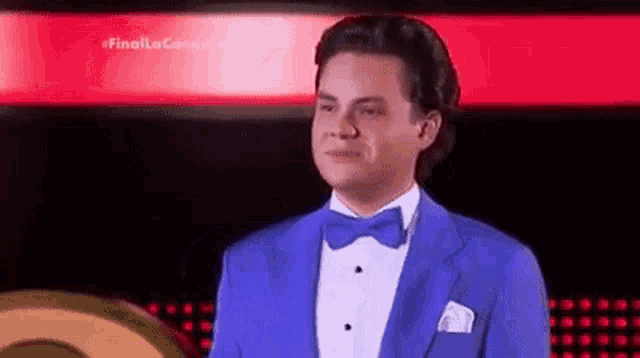a man in a blue tuxedo and bow tie is standing in front of a red background .