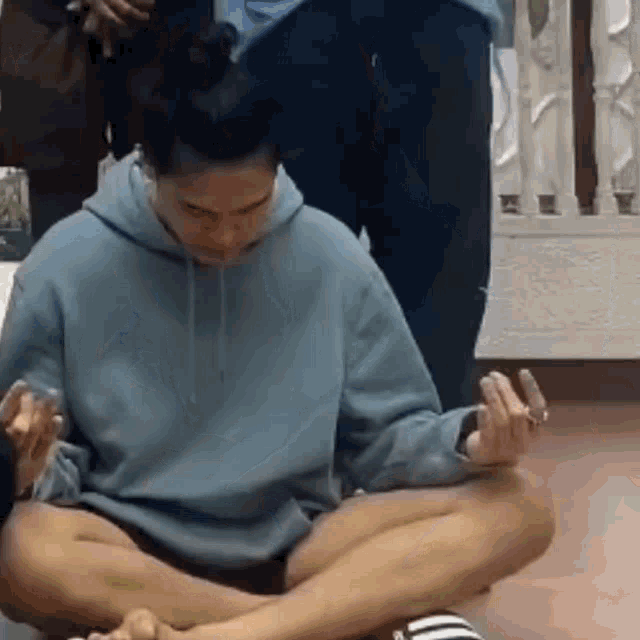 a woman in a blue hoodie is sitting on the floor with her legs crossed and her hands folded .
