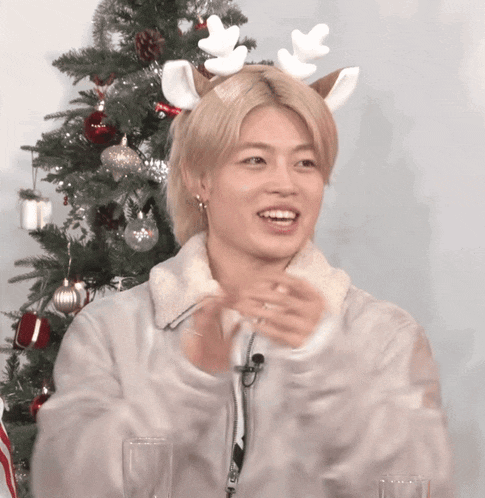 a young man wearing a reindeer antlers headband is standing in front of a christmas tree