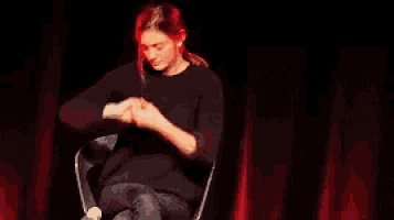 a woman with red hair is sitting in a chair with her arms outstretched in front of a red curtain .