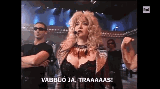 a woman with blonde hair and a microphone stands in front of a group of men and says " vabbuo ja traaaaas "