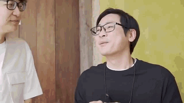 a man wearing glasses is talking to another man while standing next to a wooden wall .