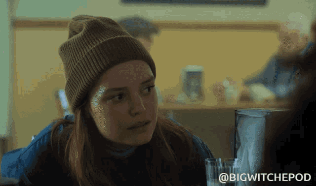 a woman wearing a beanie sits at a table with a glass of water in front of her and the words bigwitchepod