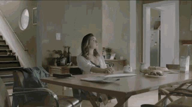 a woman sits at a table in a living room looking up