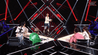 a group of people are sitting at a table in front of a stage that has a x on it