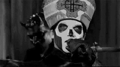 a black and white photo of a man wearing a skull mask and a priest 's hat pointing at the camera .
