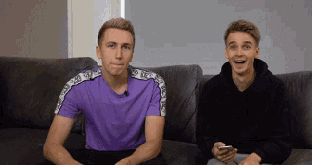 two men are sitting on a couch one is wearing a purple shirt and the other is wearing a black sweatshirt