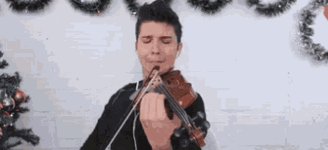 a man is playing a violin in front of a christmas tree with his eyes closed .