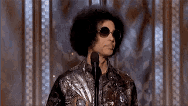 a man with an afro and sunglasses is speaking into a microphone .