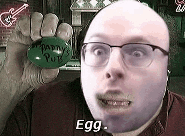 a bald man wearing glasses is holding a green egg that says `` paddy 's pub '' .