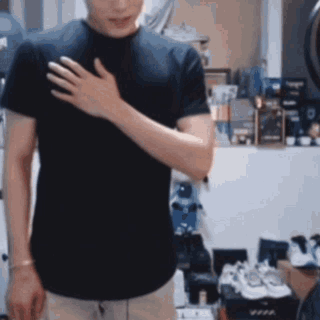 a man in a black t-shirt is standing in a room with his hands on his chest .