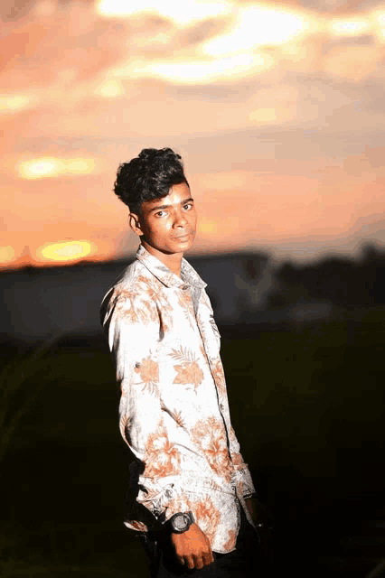 a young man in a floral shirt is standing in a field at sunset