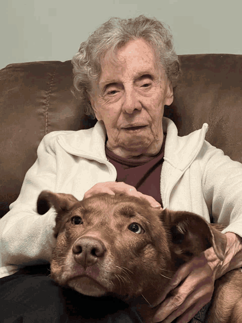 an elderly woman sits on a couch with a brown dog on her lap