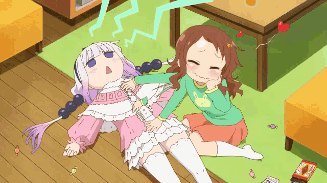 a girl in a green shirt with a cat on it sits next to a girl laying on the floor