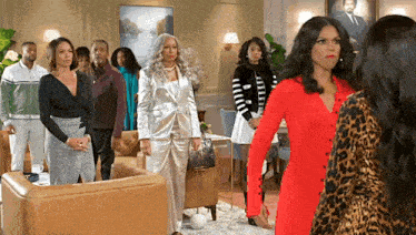 a group of women are standing in a living room . one of the women is wearing a red dress .