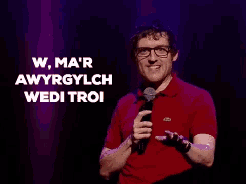 a man wearing glasses and a red shirt is holding a microphone and pointing at the camera .