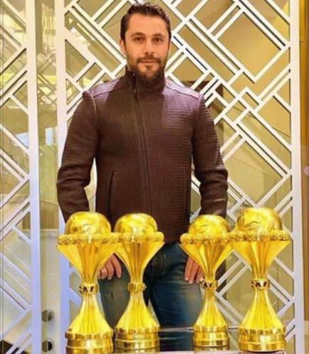 a man in a brown jacket stands next to a row of golden trophies